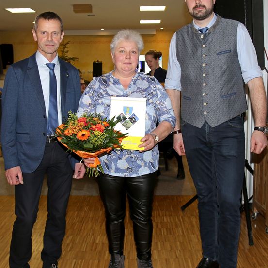 Drei Menschen stehen zusammen in einem Innenraum, eine Frau hält einen Blumenstrauß und ein Zertifikat. Der Mann auf ihrer linken Seite trägt einen Anzug, und der Mann auf ihrer rechten Seite trägt eine Weste und Jeans. Hinter ihnen geht eine Frau, und verschiedene Lichter und Gegenstände sind im Hintergrund.