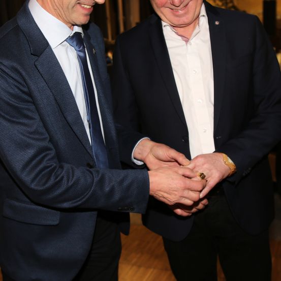 Zwei Männer in formeller Kleidung stehen lächelnd nebeneinander. Einer gibt dem anderen einen Ring. Sie tragen beide Anzüge, einer mit Krawatte und der andere mit weißem Hemd.