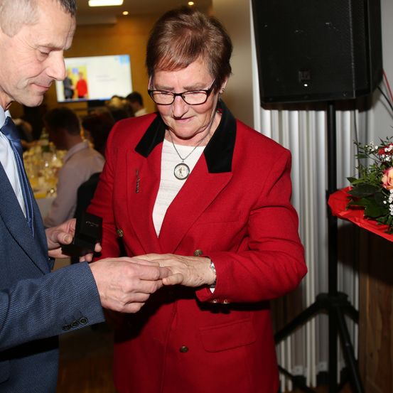 Ein Mann überreicht einer älteren Frau in einem roten Jackett eine Auszeichnung und hält eine Schachtel. Dahinter ist ein Lautsprecher zu sehen, und ein Bildschirm zeigt ein Bild an. Im Hintergrund sitzen Leute an Tischen, und ein Blumenstrauß ruht auf einer roten Oberfläche.