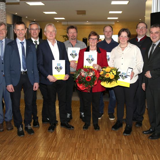 Eine Gruppe von Menschen steht in einem Raum und hält Urkunden und Blumen. Zwei Frauen in der Mitte halten Blumensträuße. Eine Frau in der Mitte hält eine Urkunde. Sie alle lächeln.