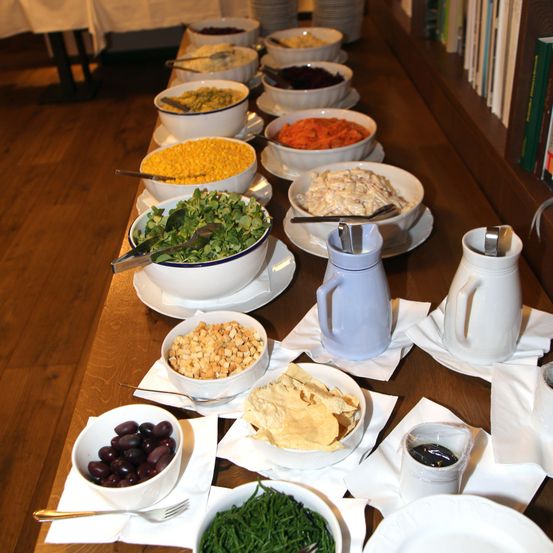 Ein Holztisch mit verschiedenen Schüsseln darauf, einschließlich Salate, Mais, Karotten, Oliven und Pommes. Die Schüsseln werden von Servietten, Löffeln und Gabeln begleitet. Zwei Gläser und zwei weiße Teller sind ebenfalls vorhanden.
