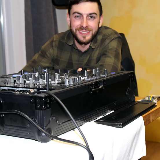 Ein Mann mit Bart sitzt vor einem DJ-Mixer auf einem Tisch. Er lächelt und scheint zu arbeiten.