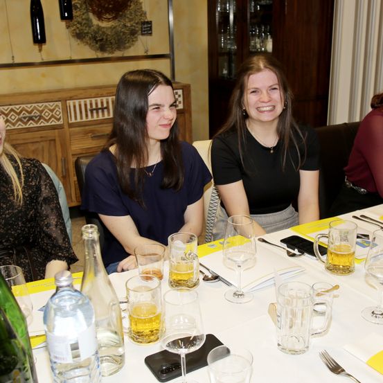 Drei Frauen sitzen an einem Tisch, lächeln und schauen sich an. Der Tisch hat Biergläser, Weingläser und eine Flasche Wasser. Dahinter steht ein Holzschrank und ein Regal mit Flaschen.