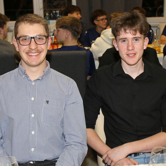 Zwei junge Männer lächeln und sitzen an einem Tisch, wahrscheinlich bei einer sozialen Veranstaltung. Der Mann auf der linken Seite trägt eine Brille und ein kariertes Hemd. Hinter ihnen sitzen mehrere Personen an einem Tisch, möglicherweise bei einer Mahlzeit.