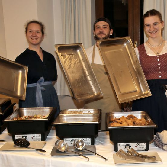 Drei Personen posieren mit Metallschalen in einer Küche, einer hält einen Schöpflöffel, ein anderer eine Pfannenwender und der dritte eine Servierschale. Auf dem Tresen stehen Schalen mit Essen.