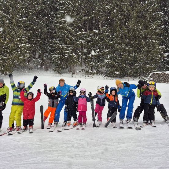 Eine Gruppe von Kindern und Erwachsenen posiert für ein Foto auf einer verschneiten Piste mit immergrünen Bäumen im Hintergrund.