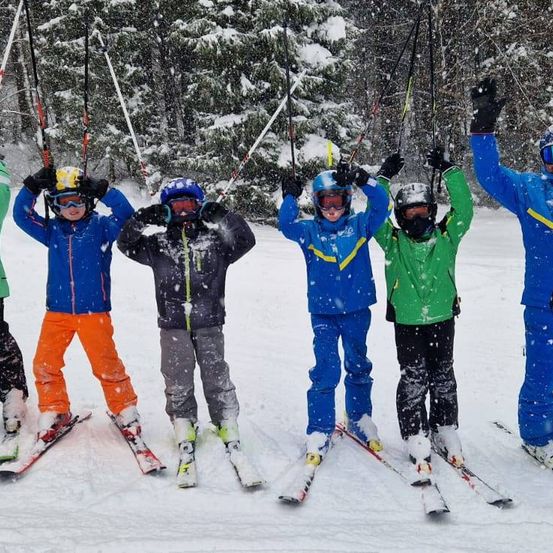 Sechs Kinder auf Skiern stehen im Schnee mit Skistöcken, Helmen und Schutzbrillen. Sie tragen bunte Jacken und Hosen. Bäume sind hinter ihnen, und es schneit.