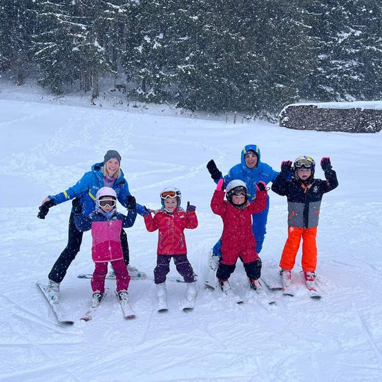 Eine Familie von sechs Personen lächelt im Schnee, alle in Winterkleidung und Skiern. Sie stehen auf einer verschneiten Piste, mit Bäumen im Hintergrund, die mit Schnee bedeckt sind.