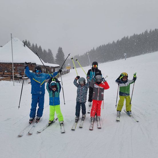 Fünf Kinder stehen im Schnee vor einer Skihütte und sind am Skifahren. Sie tragen Winterkleidung und halten Skistöcke in der Hand.