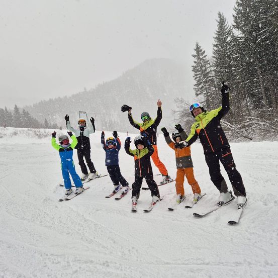 Eine Gruppe von Kindern und Erwachsenen schippert auf einem verschneiten Berg, alle tragen Skiausrüstung und Helme, sie posieren für ein Foto mit erhobenen Händen.