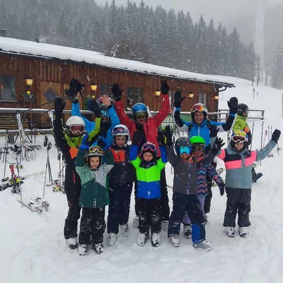 Eine Gruppe junger Skifahrer posiert für ein Foto im Schnee, mit Helmen und Schutzbrillen. Sie stehen vor einem Gebäude mit schneebedecktem Dach und Bäumen.