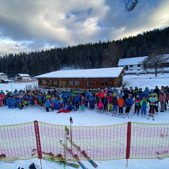 Eine Gruppe von Skifahrern und Snowboardern posiert für ein Foto vor einem Gebäude, umgeben von einer verschneiten Landschaft und Bäumen.