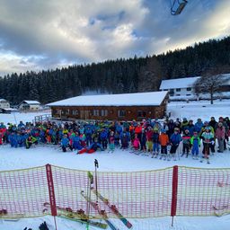 Eine Gruppe von Skifahrern und Snowboardern posiert für ein Foto vor einem Gebäude, umgeben von einer verschneiten Landschaft und Bäumen.