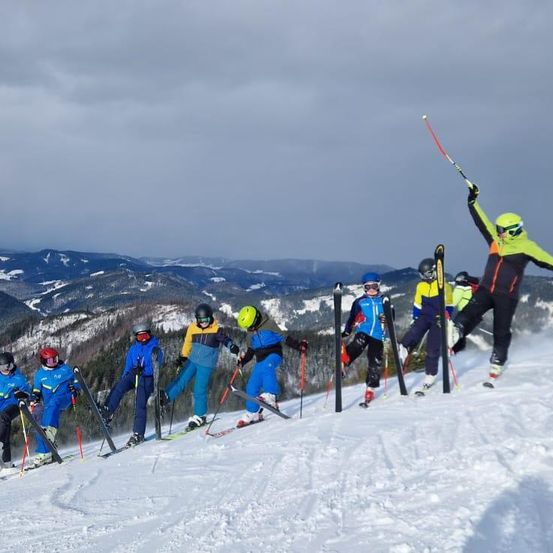 Eine Gruppe von Skifahrern ist auf einem verschneiten Berghang mit bewölktem Himmel. Sie tragen farbenfrohe Skianzüge und Helme, einige mit Skistöcken. Ein Skifahrer springt in die Luft mit erhobenen Skistöcken.