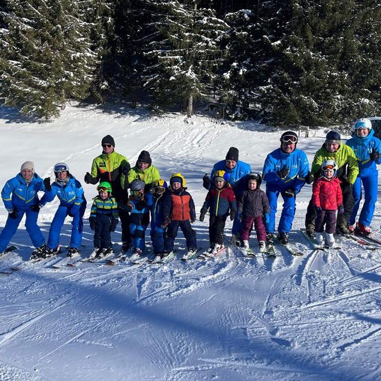 Eine Gruppe von Skifahrern in Skiausrüstung steht auf einem verschneiten Berg mit immergrünen Bäumen im Hintergrund.