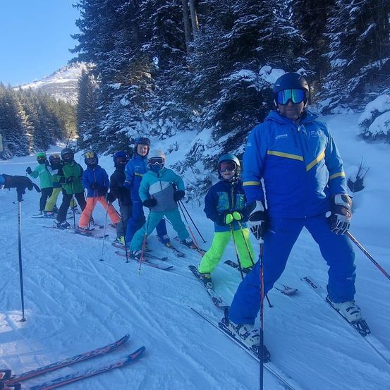 Eine Gruppe Skifahrer, darunter Kinder und ein Ausbilder, steht auf einer verschneiten Piste. Sie sind alle in Skiausrüstung gekleidet, einschließlich Helmen und Handschuhen.