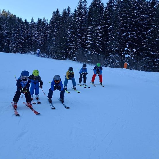 Eine Gruppe von Kindern fährt auf einem verschneiten Hang Ski, trägt Helme und Skianzüge. Der verschneite Wald im Hintergrund ist schneebedeckt.