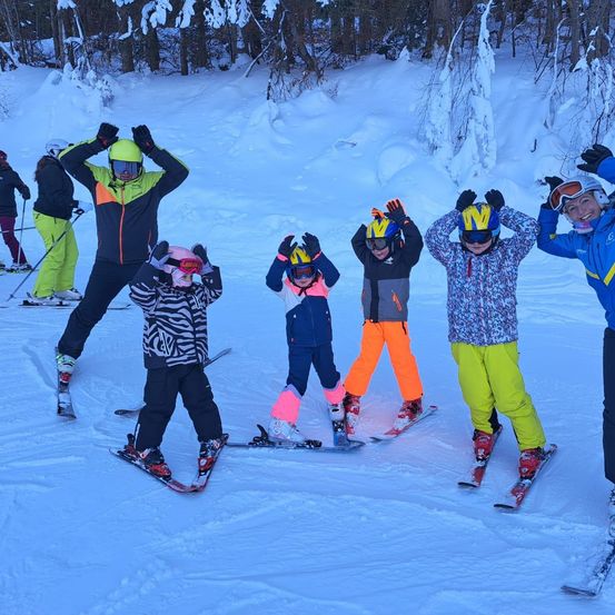 Eine Gruppe von Skifahrern, einschließlich Kindern und Erwachsenen, posiert für ein Foto im Schnee. Sie tragen alle Helme und Brillen. Hinter ihnen fahren zwei Personen die Piste hinunter.