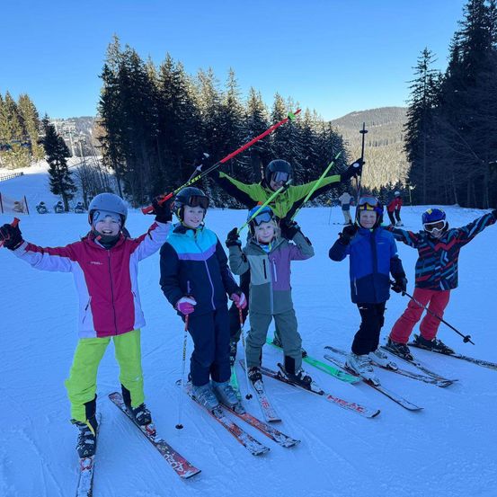 Eine Gruppe von Kindern in Skiausrüstung steht auf einem verschneiten Hang und lächelt in die Kamera. Dahinter springt eine Person mit erhobenen Skistöcken. Im Hintergrund sind Bäume und ein Berg zu sehen.