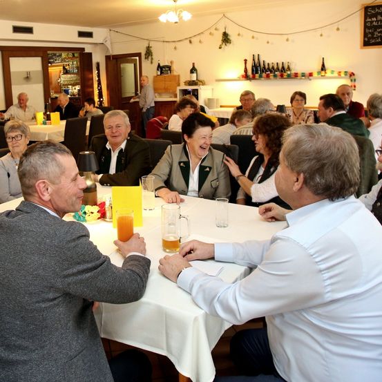 Eine Gruppe älterer Menschen sitzt um einen Tisch herum und unterhält sich. Einige lachen. Auf dem Tisch stehen Getränke und eine Speisekarte. Dahinter stehen Flaschen auf einem Regal und eine Tafel.