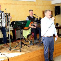 Eine Gruppe von Musikern tritt auf einer Bühne auf. Ein Mann spielt Akkordeon, ein anderer Gitarre und ein dritter Mann singt in ein Mikrofon. Hinter ihnen sind verschiedene Musikinstrumente und Ausrüstung aufgebaut.