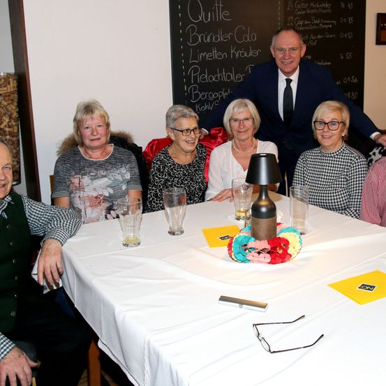 Eine Gruppe älterer Menschen sitzt lächelnd um einen Tisch. Auf dem Tisch befinden sich Gläser und eine Flasche. Hinter ihnen steht ein Mann im Anzug, der lächelt und seine Hand auf die Schulter einer Frau legt.