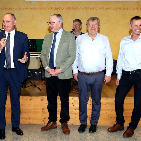 Fünf Männer in Anzügen und Krawatten stehen lächelnd auf der Bühne. Der Mann auf der linken Seite hält ein Mikrofon. Hinter ihnen spricht ein Mann in ein Mikrofon auf einem Ständer.
