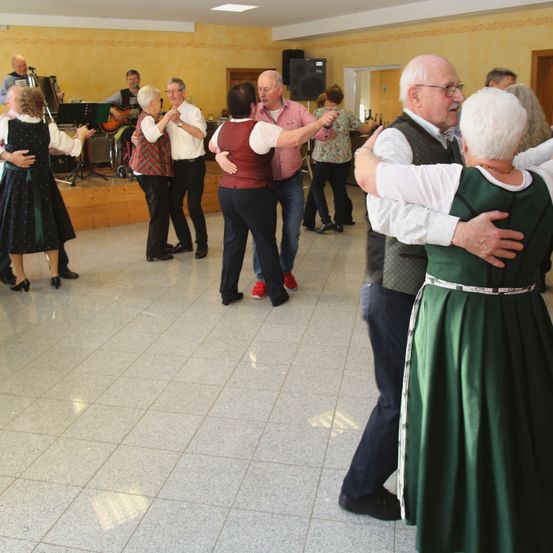 Eine Gruppe älterer Erwachsener tanzt in einem Saal mit gefliesten Böden. Einige tragen traditionelle Kleidung. Hinter ihnen spielen Musiker auf einer Bühne mit einem Lautsprecher.