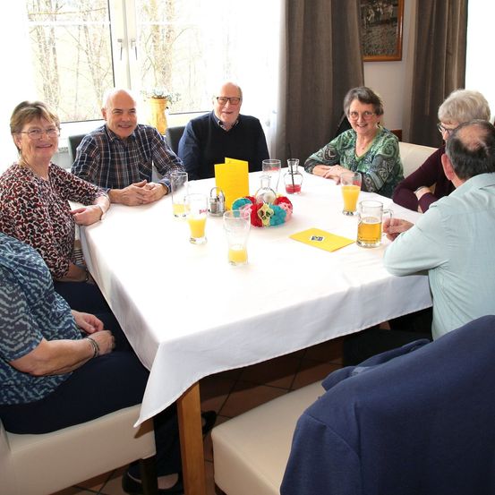 Eine Gruppe Erwachsener sitzt um einen Tisch herum, lächelt und schaut in die Kamera. Der Tisch ist mit einer weißen Tischdecke bedeckt. Auf dem Tisch stehen Gläser mit Orangensaft und eine Speisekarte. Auf dem Tisch befinden sich auch dekorative Gegenstände. Hinter ihnen ist ein Fenster mit Blick auf Bäume.