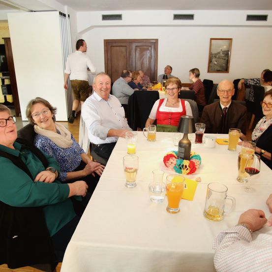 Eine Gruppe von Menschen sitzt an einem Tisch mit Getränken und Essen und lächelt. Dahinter geht eine Person am Tisch vorbei.
