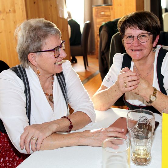Zwei Frauen mit Brillen und Halsketten sitzen auf Stühlen, lachen und unterhalten sich. Sie sitzen vor einem Tisch mit Gläsern. Dahinter sitzt eine Person auf einem Stuhl.