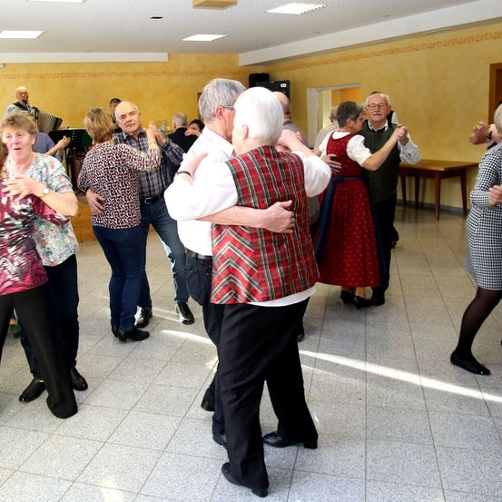 Eine Gruppe älterer Menschen tanzt in einem Raum mit gelben Wänden, der von Deckenleuchten beleuchtet wird. Ein Mann spielt Akkordeon an der Wand, während andere stehen oder auf Stühlen sitzen.