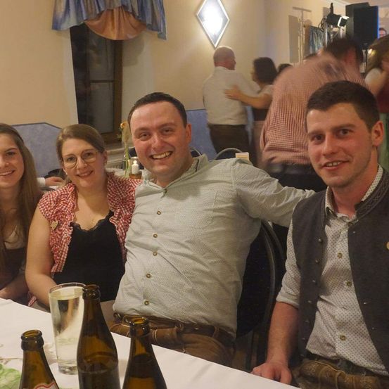 Drei Personen sitzen mit Bierflaschen an einem Tisch und lächeln. Eine Frau und ein Mann sitzen, wobei der Mann die Frau umarmt. Ein Mann in traditioneller deutscher Kleidung sitzt neben ihnen. Im Hintergrund tanzen Menschen.