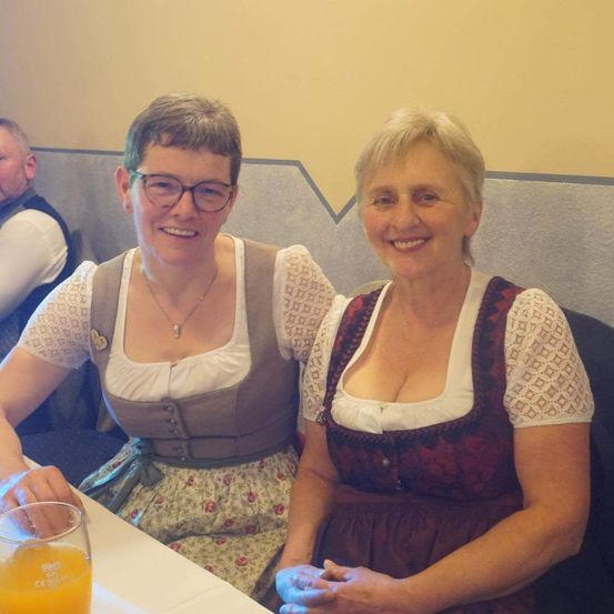 Zwei lächelnde Frauen, eine mit Brille, sitzen an einem Tisch. Sie tragen traditionelle Kleider und ein Mann sitzt hinter ihnen. Ein Glas Orangensaft steht auf dem Tisch.