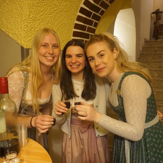 Drei Frauen in traditioneller Kleidung posieren mit Getränken in einem Raum mit gelber Wand und Treppe.