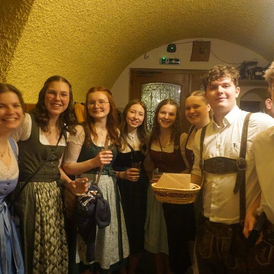 Eine Gruppe von Menschen in traditioneller Kleidung posiert für ein Foto. Sie halten Getränke und einen Korb mit Servietten. Der Hintergrund zeigt eine gelbe Wand und eine Holztür.