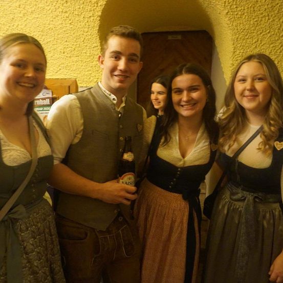 Vier Menschen in traditioneller bayerischer Kleidung lächeln für ein Foto. Ein Mann hält eine Bierflasche. Hinter ihnen ist eine teilweise sichtbare gelbe Wand mit einem Schild zu sehen.