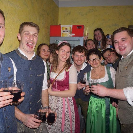 Eine Gruppe von Menschen in traditioneller bayerischer Kleidung posiert für ein Foto, einige mit Getränken in den Händen, vor einem Kaiser-Bierkühlschrank.