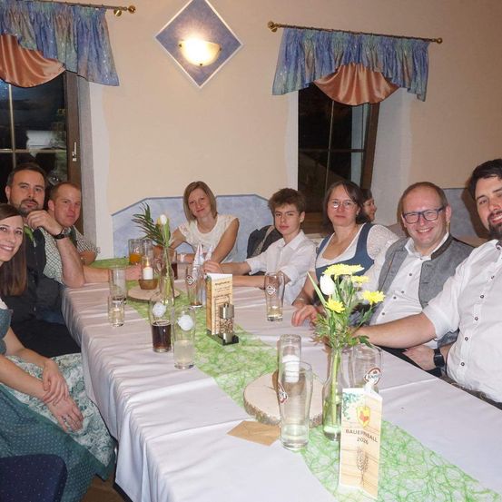 Eine Gruppe von Menschen sitzt um einen Tisch und lächelt für ein Foto. Der Tisch ist mit einem weißen Tuch bedeckt und hat eine Blumenarrangement, Gläser und Menükarten. Der Raum hat beige Wände mit Vorhängen und einer Lampe.