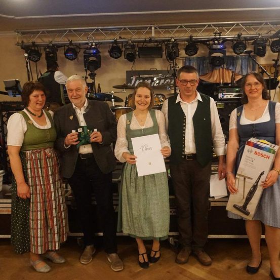 Fünf Personen stehen zusammen auf einer Bühne mit einem Mikrofon und lächeln für ein Foto. Eine Frau hält ein Zertifikat. Ein Schlagzeug und andere Musikinstrumente stehen hinter ihnen.