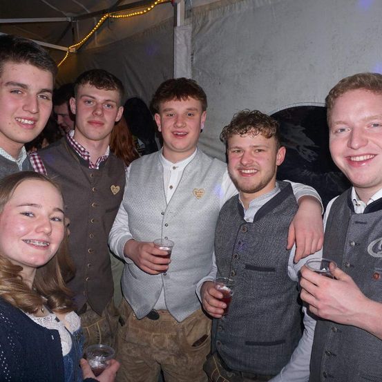 Eine Gruppe junger Leute in traditioneller deutscher Kleidung, lächelnd und Getränke in der Hand bei einer Veranstaltung.