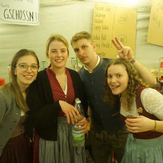 Vier junge Erwachsene posieren für ein Foto bei einer Veranstaltung, in traditioneller bayerischer Kleidung. Dahinter zeigt ein gelbes Schild Getränkeoptionen an.