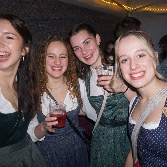 Vier Frauen lächeln und posieren für ein Foto, wahrscheinlich auf einem Festival. Sie tragen traditionelle grüne Kleider und halten Getränke in den Händen. Eine von ihnen trägt eine Halskette. Hinter ihnen ist ein verschwommener Hintergrund von Menschen zu sehen, und ein Zelt mit Lichtern ist sichtbar.