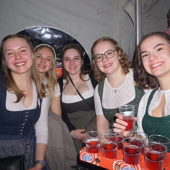 Fünf junge Frauen lächeln für ein Foto auf einem Festival, eine hält einen Tablett mit Getränken. Die Frau auf der rechten Seite trägt eine Brille und eine Halskette.