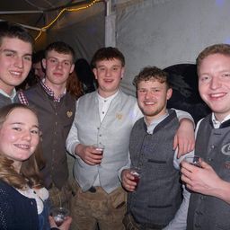 Eine Gruppe junger Männer und Frauen lächelt und posiert für ein Foto, während sie Gläser mit Getränken in den Händen halten. Sie tragen traditionelle Kleidung. Hinter ihnen ist ein weißes Zelt zu sehen.