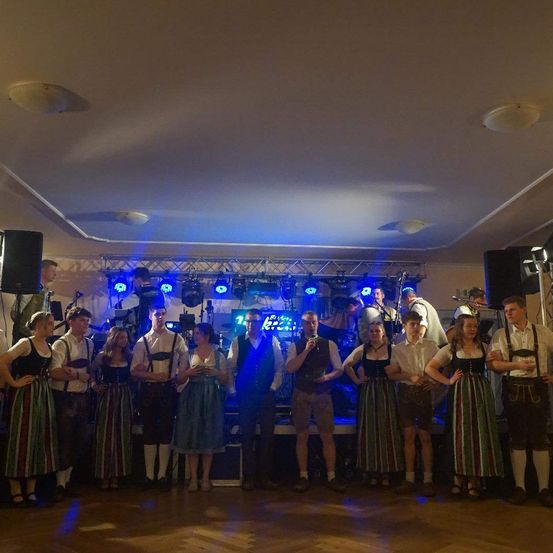 Eine Gruppe von Menschen in traditioneller deutscher Kleidung steht auf einer Bühne, möglicherweise bei einer Feier. Sie werden von blauen Bühnenlichtern beleuchtet.