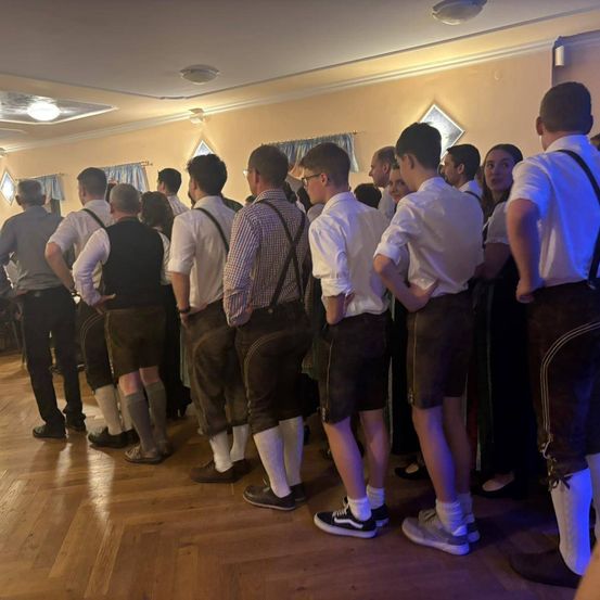 Eine Gruppe von Männern und Frauen steht in einem Raum und trägt traditionelle Kleidung. Sie schauen in die gleiche Richtung.