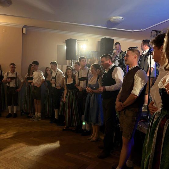 Eine Gruppe von Menschen in traditioneller bayerischer Kleidung, die in einem Raum mit Holzboden stehen, einige mit Getränken in der Hand. Ein Mikrofon auf einem Ständer steht vor ihnen.