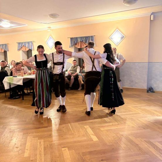 Drei Menschen tanzen in einem Ballsaal, zwei Frauen und ein Mann. Sie sind von einer Menschenmenge umgeben, die an Tischen sitzt.