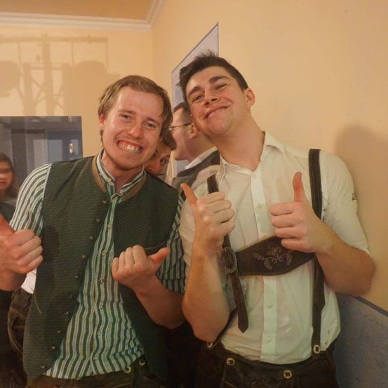 Zwei Männer in traditioneller deutscher Kleidung lächeln und geben Daumen hoch. Einer trägt eine grüne Weste, der andere ein weißes Hemd und eine Hose mit Hosenträgern. Sie stehen neben einer gelben Wand.